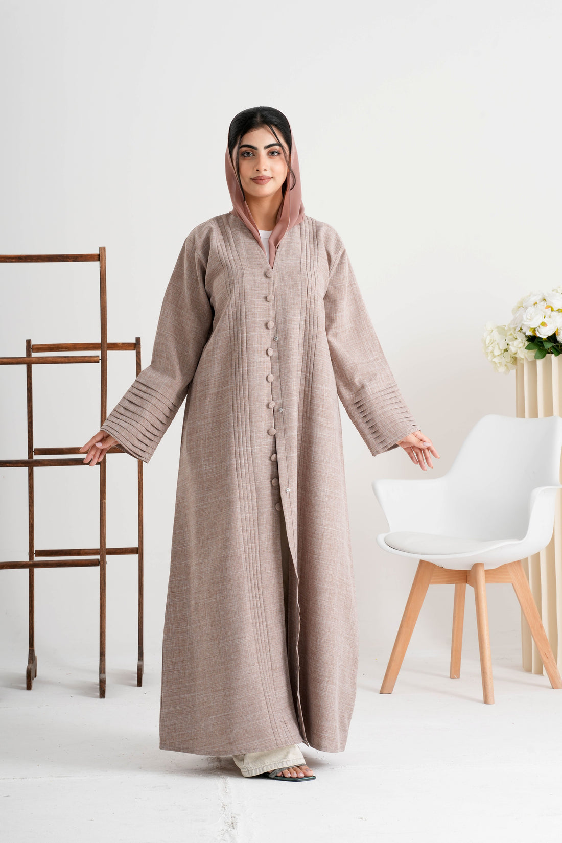 Woman wearing a long beige dress in a minimalistic room with white walls and a white chair.